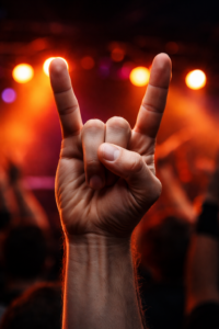 Close-up of a raised human hand making the Satan’s Greeting hand gesture at a live concert, with stage lights and a blurred crowd in the background