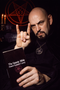 Portrait of Anton LaVey holding The Satanic Bible while making the Satan’s Greeting hand gesture, with occult symbols and candlelight in the background