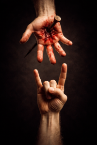 Symbolic image showing a wounded human hand pierced by a nail above another hand making the Satan’s Greeting gesture against a dark background